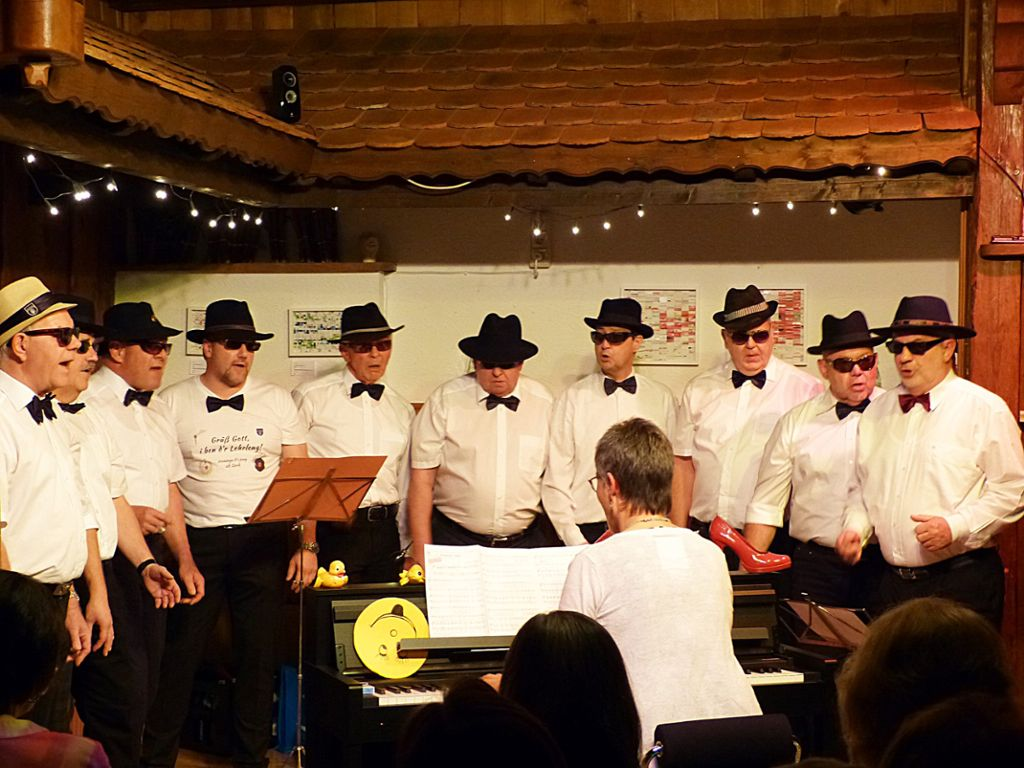 10 alte Männer mit weißem Hemd und Fliege und Hut singen. Im Vordergrund eine Frau am Klavier