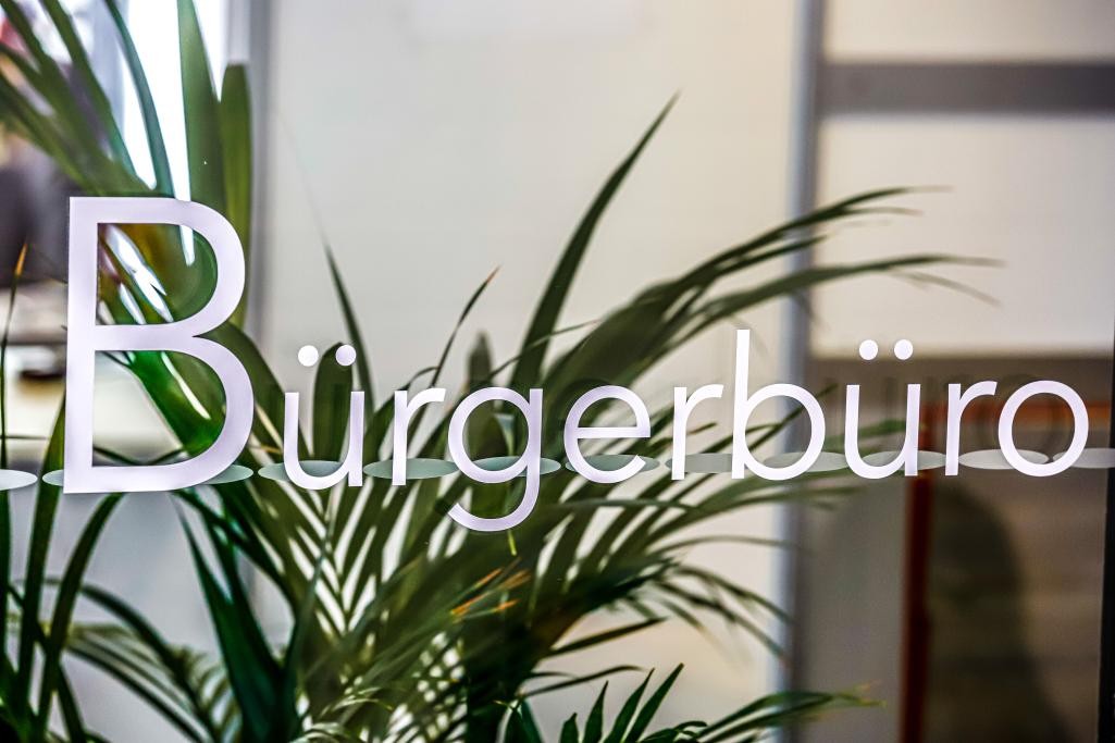 Arbeitsplatz im Bürgerbüro im Rathaus Schriftzug Bürgerbüro auf Glasscheibe, dahinter Pflanze