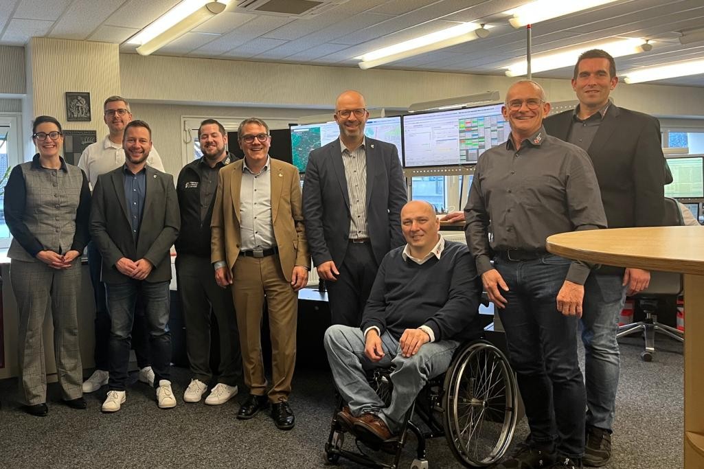 Eine Gruppe von Personen in einnem hellen Raum mit mehreren Bildschirmen im Hintergrund