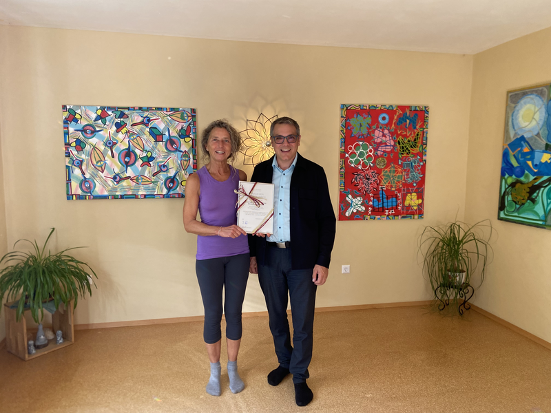 Mann und Frau stehen mit einer Urkunde im Yogaraum,an der Wand hängen bunte Bilder