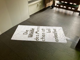 Die Würde des Menschen Werk "Die Würde des Menschen" Eine weiße Fahne mit einem Schriftzug liegt auf dem Boden
