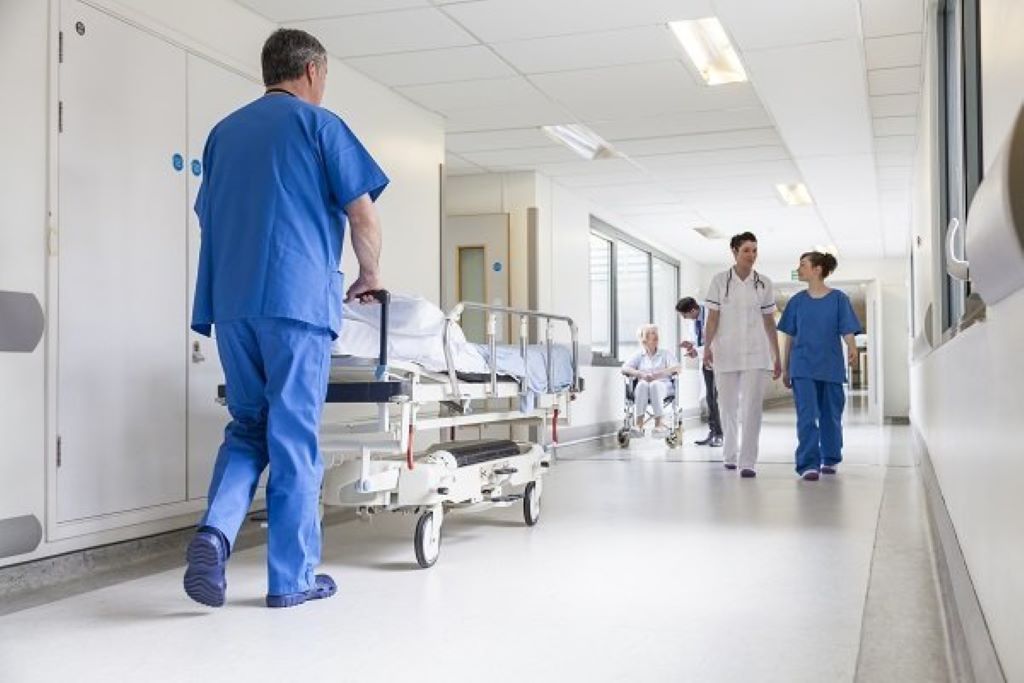 Krankenhausflur - Pfleger schiebt Krankenhausbett und Patientin im Rollstuhl sowie weiteres Pflegepersonal geht den Gang entlang