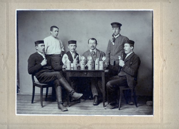 Schwarzweiss Foto von sechs Männern um einen Tisch gruppiert mit Bierkrügen
